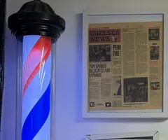 A lit red, white and blue barber pole beside a framed Chelsea News front page about the shop's move